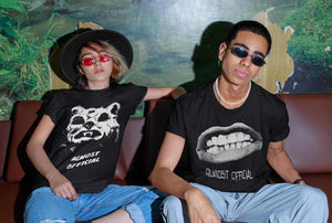 A man and a woman sitting on a couch. The woman is wearing the Almost Official Hombre Lobo shirt, and the man is wearing the Almost Official Pearly Whites shirt.