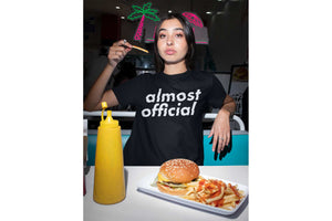 A girl wearing an Almost Official Glitch Logo t shirt while eating a burger and fries