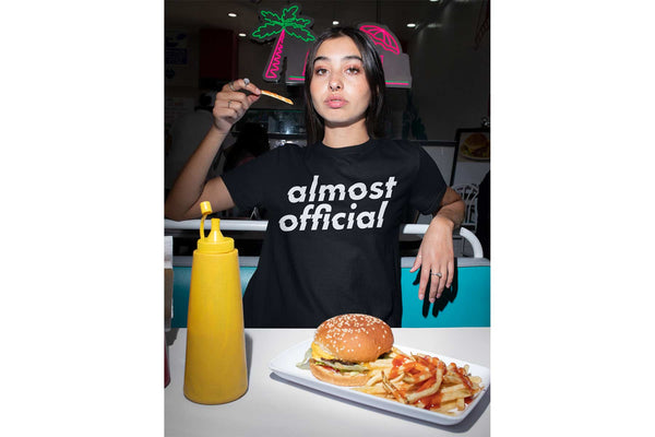 A girl wearing an Almost Official Glitch Logo t shirt while eating a burger and fries