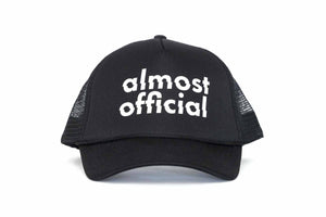 The front of a black trucker hat with the Almost Official Logo printed in white. 