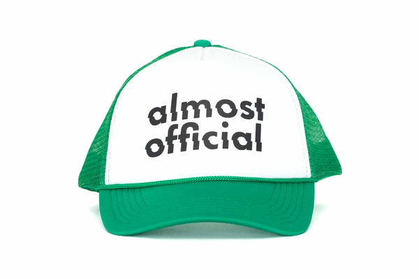 The front of a green and white trucker hat with the Almost Official Logo printed in black. 