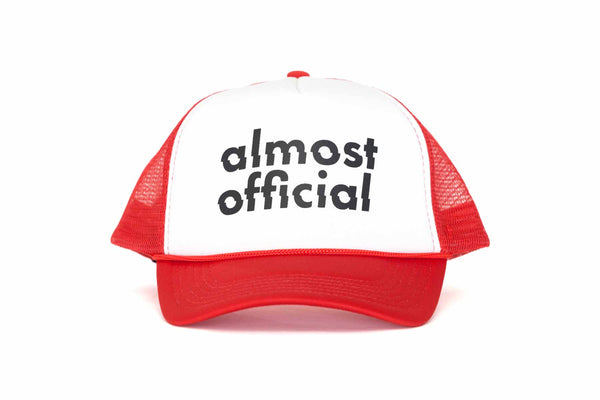 The front of a red and white trucker hat with the Almost Official Logo printed in black. 