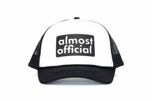 The front of a black and white trucker hat with the Almost Official black stamp logo printed on the front panel. 