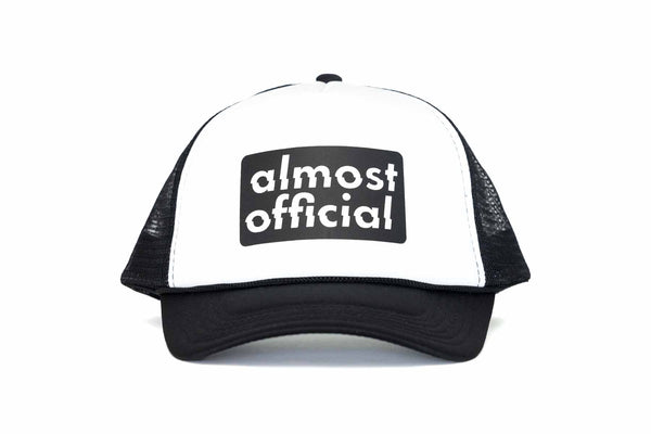 The front of a black and white trucker hat with the Almost Official black stamp logo printed on the front panel. 